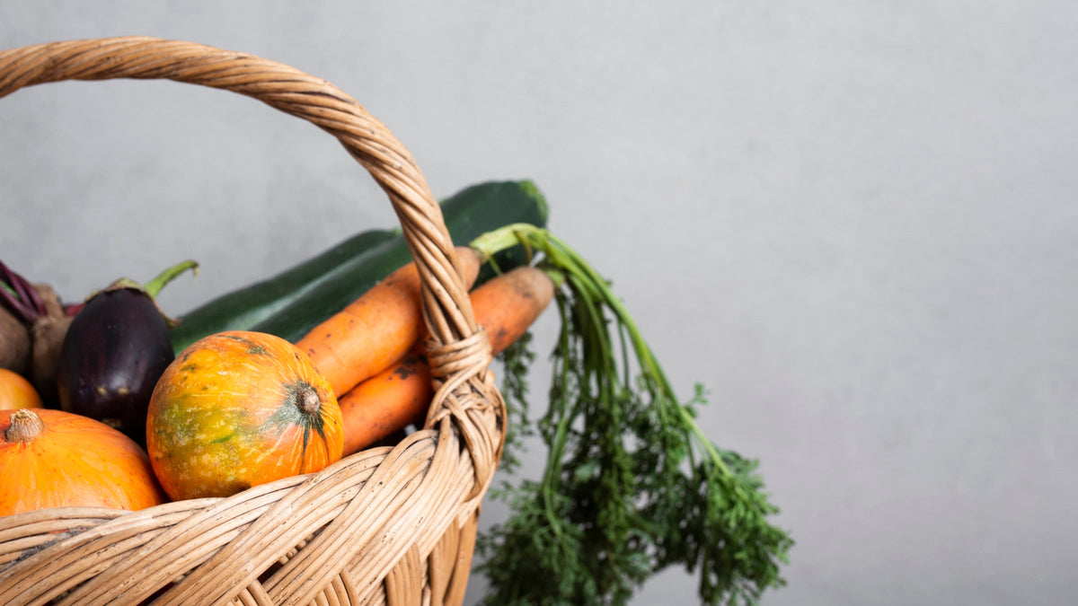 Cesta de mimbre con productos de temporada y alimentos frescos como zanahorias, calabacines, berenjenas y calabazas sobre fondo gris claro