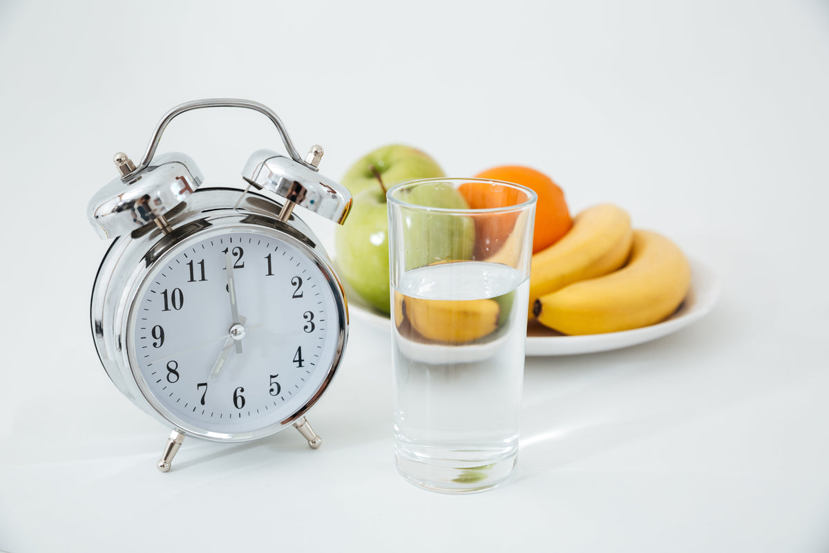 Reloj despertador junto a un vaso de agua y frutas frescas, representando el ayuno intermitente y la alimentación saludable