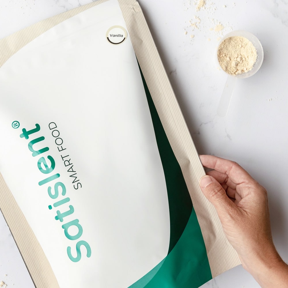 Bag of Satislent Food vanilla flavour on marble, next to a scoop of powder