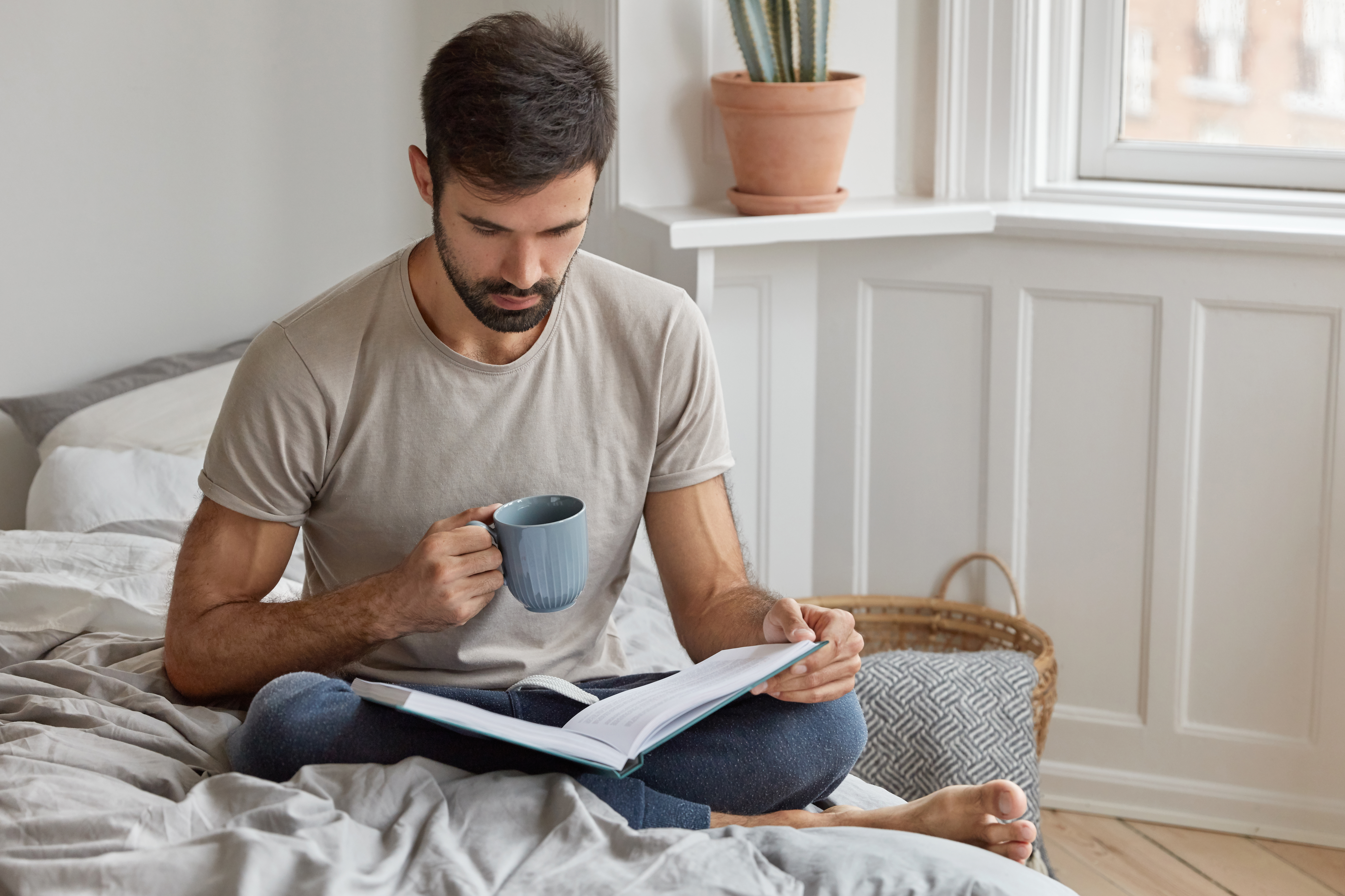 Homme assis sur un lit avec une tasse et un livre, illustrant une routine matinale calme et consciente dans le Monk Mode